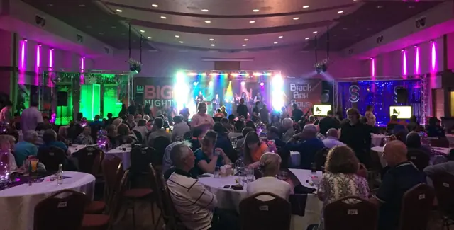 A packed function room full of tables, with coloured stage lighting at the far end