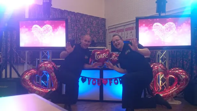 Darren and Colin at a Valentines disco with heart balloons and pink screens